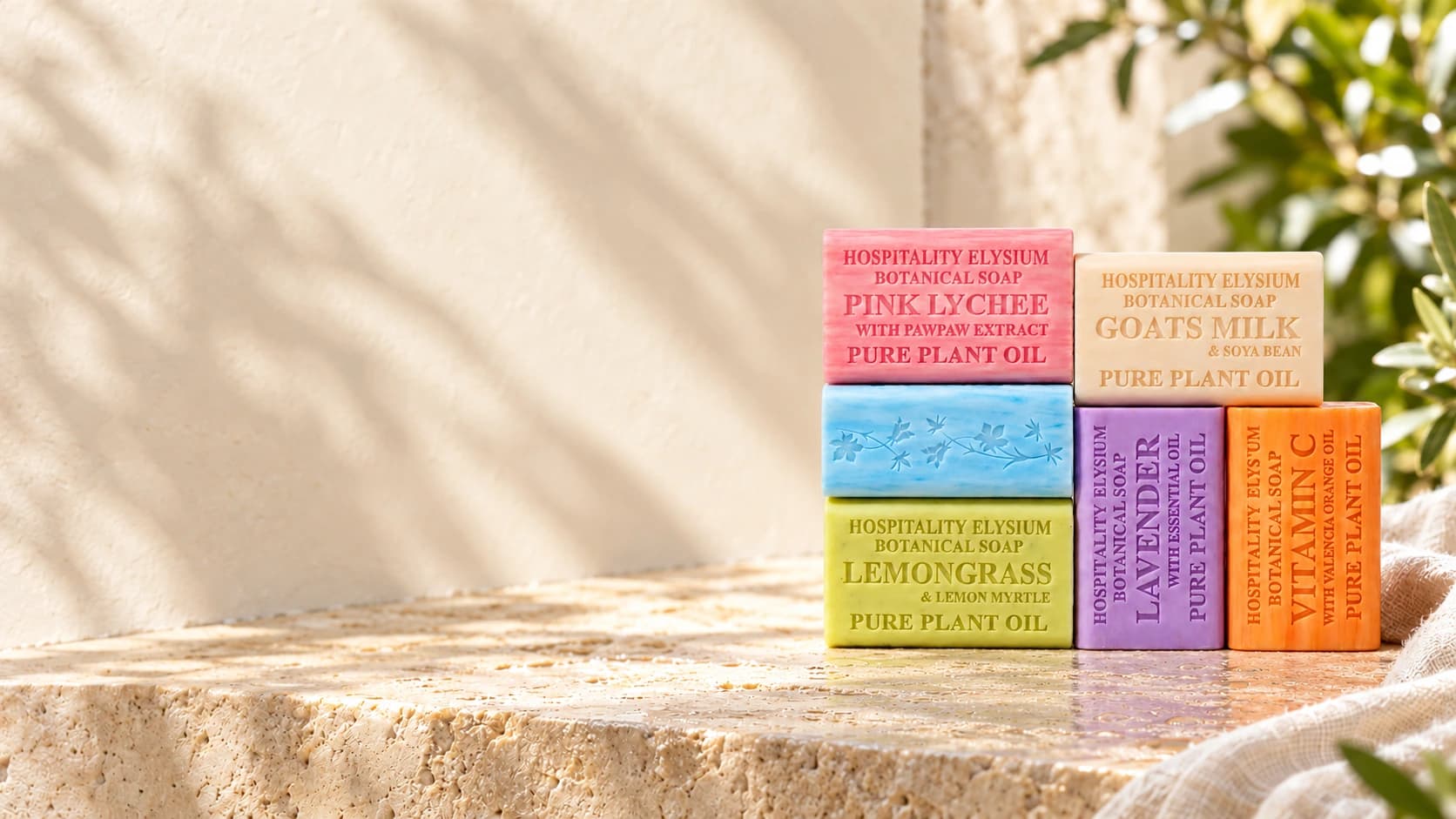 Six colourful Hospitality Elysium botanical soap bars with embossed lettering and floral detail on a sunlit stone ledge